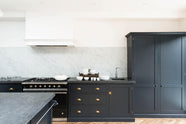 A bright and sophisticated pantry-blue Shaker kitchen features a sink and cooker run with a Belgian blue limestone worktop, a Carrara marble splashback, full-height cupboards, a canopy, and a kitchen island. Adorned with plain white ceramics, short drinking glasses, a black gas cooker, and beautiful brass fittings, it creates a functional and contemporary kitchen space.