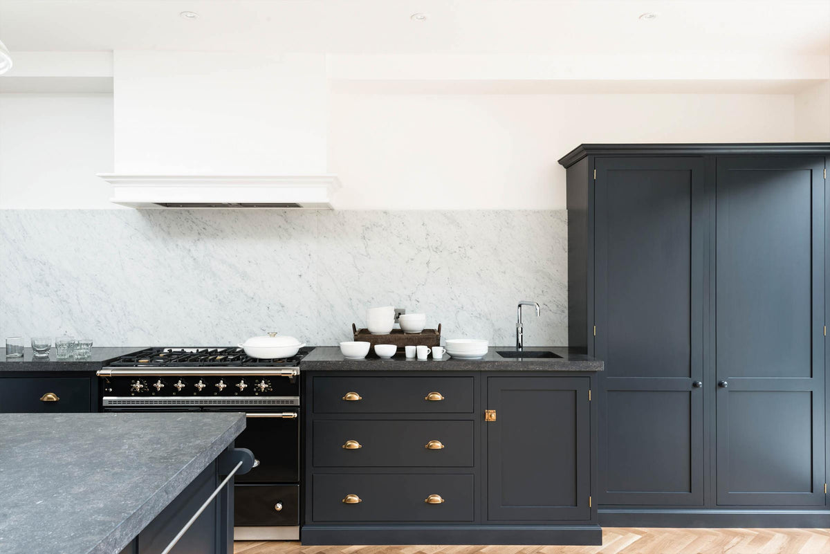 A bright and sophisticated pantry-blue Shaker kitchen features a sink and cooker run with a Belgian blue limestone worktop, a Carrara marble splashback, full-height cupboards, a canopy, and a kitchen island. Adorned with plain white ceramics, short drinking glasses, a black gas cooker, and beautiful brass fittings, it creates a functional and contemporary kitchen space.