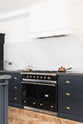 An oblique view of a bright and sophisticated pantry-blue Shaker kitchen features a sink and cooker run with a Belgian blue limestone worktop, a Carrara marble splashback, and a kitchen island. Adorned with a copper saucepan, wire baskets with fresh eggs, a black gas cooker, and beautiful brass fittings, it creates a functional and contemporary kitchen space.