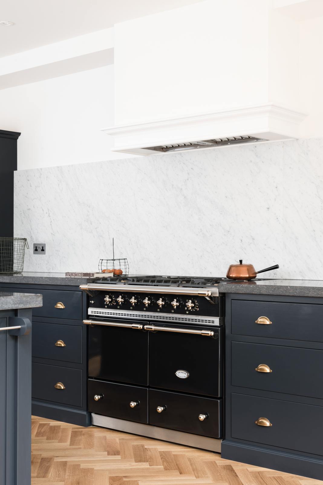 An oblique view of a bright and sophisticated pantry-blue Shaker kitchen features a sink and cooker run with a Belgian blue limestone worktop, a Carrara marble splashback, and a kitchen island. Adorned with a copper saucepan, wire baskets with fresh eggs, a black gas cooker, and beautiful brass fittings, it creates a functional and contemporary kitchen space.