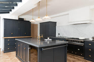 An oblique view of a bright and sophisticated pantry-blue Shaker kitchen features a sink and cooker run with a Belgian blue limestone worktop, a Carrara marble splashback, full-height cupboards, a canopy, a closed larder with a solid oak worktop, and a kitchen island. Adorned with golden-chained glass pendant lights, plain white ceramics, short drinking glasses, and beautiful brass fittings, it creates a functional and contemporary kitchen space.