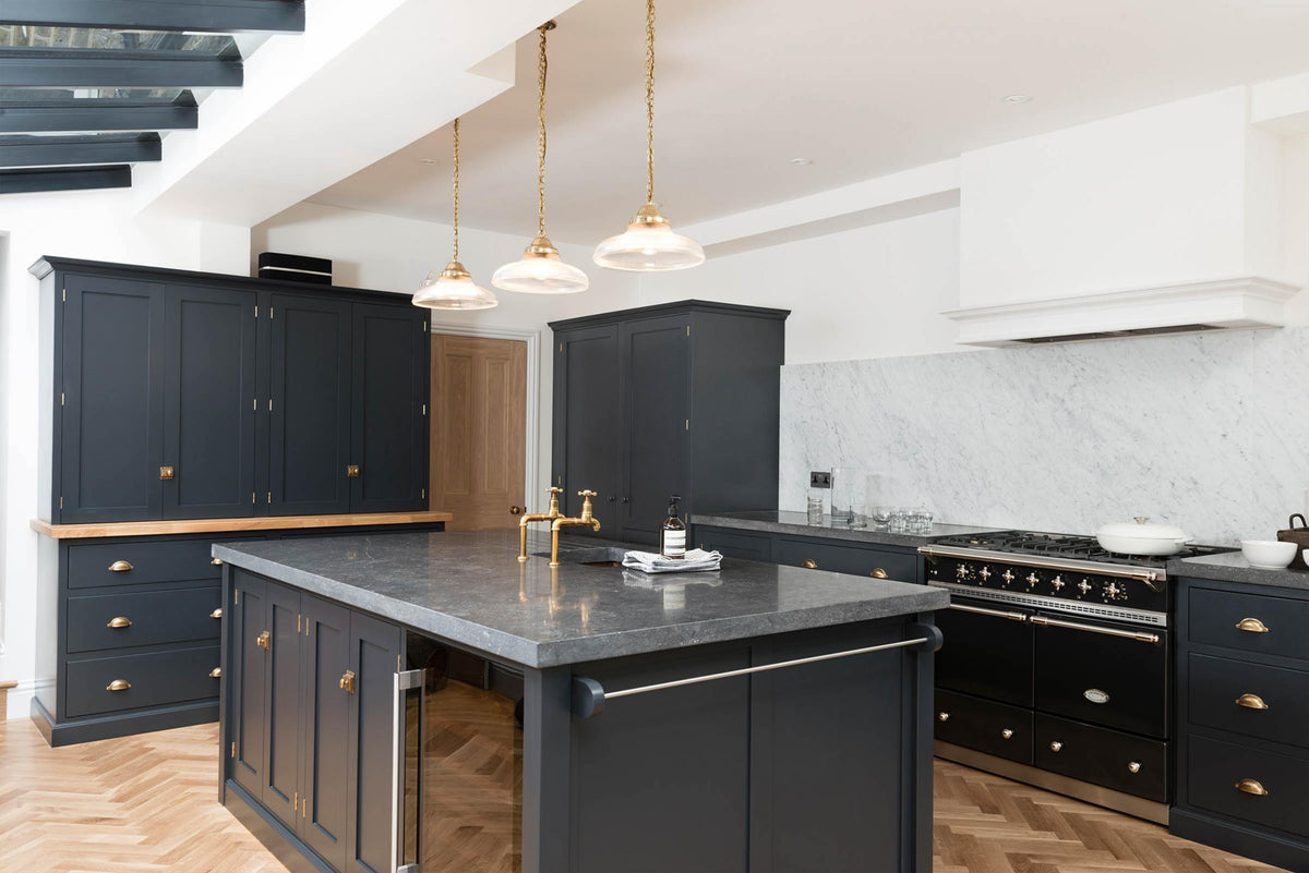An oblique view of a bright and sophisticated pantry-blue Shaker kitchen features a sink and cooker run with a Belgian blue limestone worktop, a Carrara marble splashback, full-height cupboards, a canopy, a closed larder with a solid oak worktop, and a kitchen island. Adorned with golden-chained glass pendant lights, plain white ceramics, short drinking glasses, and beautiful brass fittings, it creates a functional and contemporary kitchen space.