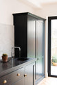 An oblique view of a bright and sophisticated pantry-blue Shaker kitchen features a sink and cooker run with a Belgian blue limestone worktop, a Carrara marble splashback and full-height cupboards. Adorned with a silver tap, a single copper sink, a cactus plant, and beautiful brass fittings, it creates a functional and contemporary kitchen space.