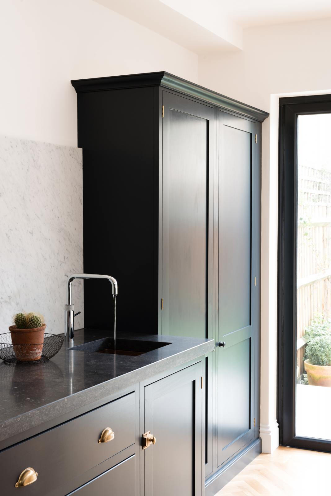 An oblique view of a bright and sophisticated pantry-blue Shaker kitchen features a sink and cooker run with a Belgian blue limestone worktop, a Carrara marble splashback and full-height cupboards. Adorned with a silver tap, a single copper sink, a cactus plant, and beautiful brass fittings, it creates a functional and contemporary kitchen space.