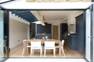 Past bifold patio doors and a dining table set, a bright and sophisticated pantry-blue Shaker kitchen features a sink and cooker run with a Belgian blue limestone worktop, a Carrara marble splashback, full-height cupboards, a canopy, a closed larder with a solid oak worktop, and a kitchen island. Adorned with golden-chained glass pendant lights and plain white ceramics, creates a functional and contemporary kitchen space.