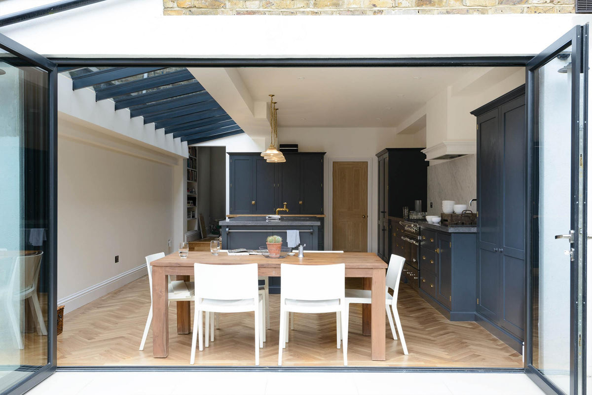 Past bifold patio doors and a dining table set, a bright and sophisticated pantry-blue Shaker kitchen features a sink and cooker run with a Belgian blue limestone worktop, a Carrara marble splashback, full-height cupboards, a canopy, a closed larder with a solid oak worktop, and a kitchen island. Adorned with golden-chained glass pendant lights and plain white ceramics, creates a functional and contemporary kitchen space.