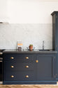 A corner of an oblique view of a bright and sophisticated pantry-blue Shaker kitchen features a sink and cooker run with a Belgian blue limestone worktop, a Carrara marble splashback, and full-height cupboards.  Adorned with a silver tap, a single copper sink, a cactus plant, and beautiful brass fittings, it creates a functional and contemporary kitchen space.