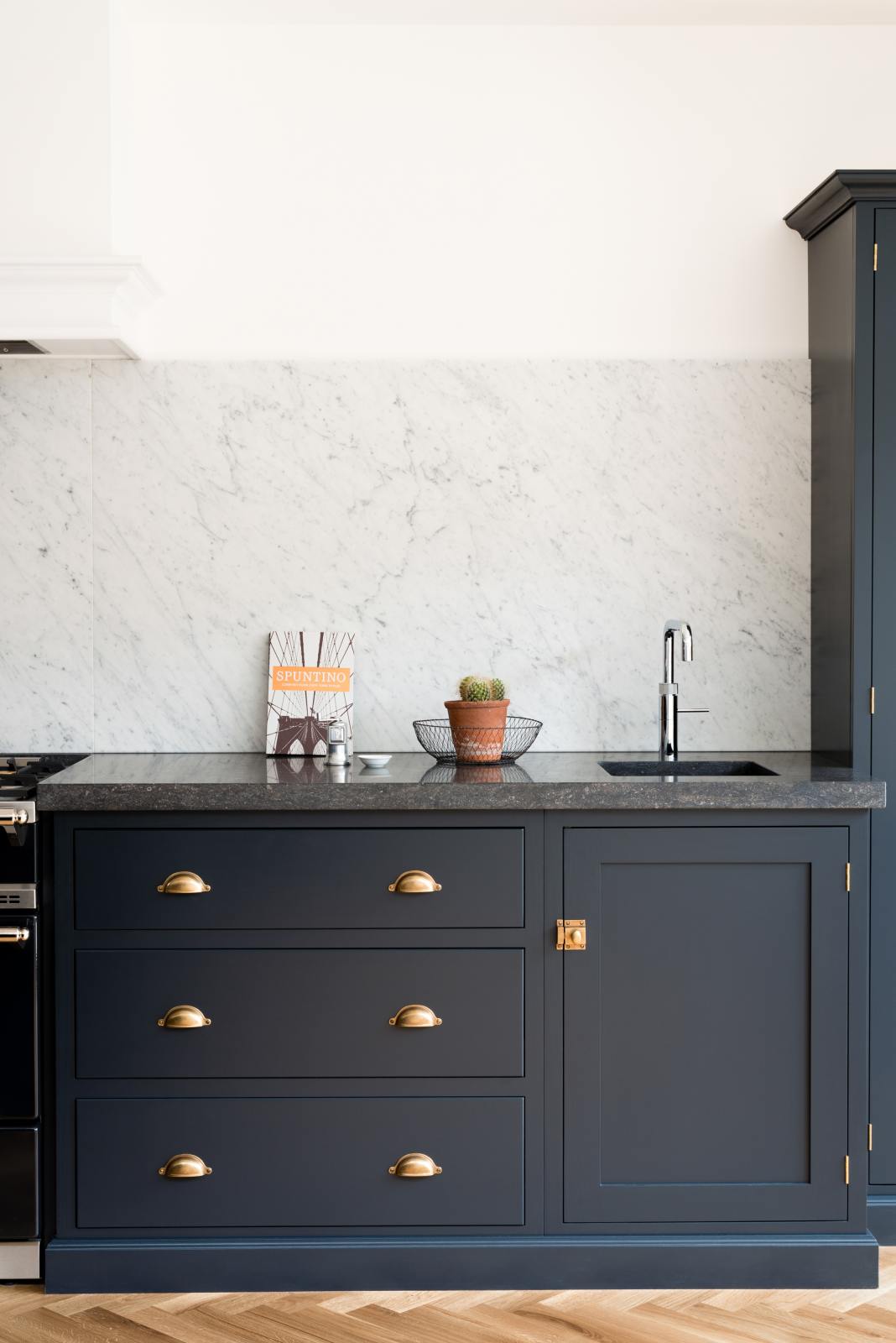 A corner of an oblique view of a bright and sophisticated pantry-blue Shaker kitchen features a sink and cooker run with a Belgian blue limestone worktop, a Carrara marble splashback, and full-height cupboards.  Adorned with a silver tap, a single copper sink, a cactus plant, and beautiful brass fittings, it creates a functional and contemporary kitchen space.