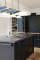 An oblique view of a bright and sophisticated pantry-blue Shaker kitchen features a sink and cooker run with a Belgian blue limestone worktop, a Carrara marble splashback, a closed larder with a solid oak worktop, and a kitchen island. Adorned with colourful books, a single copper sink with aged brass Mayan taps, and beautiful brass fittings, it creates a functional and contemporary kitchen space.