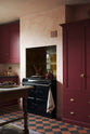 An oblique view of a warm and simply gorgeous refectory-red Shaker kitchen features a sink run with a black granite worktop, a wall cupboard, a pantry cupboard, and a gas stove in a chimney breast with Sepia Lace market splashback tiles. Beautifully decorated with vintage furniture, artisan ceramics, and a matching red teapot, it creates a humble and endearing space.