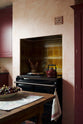 An oblique view of a warm and simply gorgeous refectory-red Shaker kitchen features a sink run with a black granite worktop, a wall cupboard, a pantry cupboard, and a gas stove in a chimney breast with Sepia Lace market splashback tiles. Beautifully decorated with vintage furniture, artisan ceramics, and a matching red teapot, it creates a humble and endearing space.