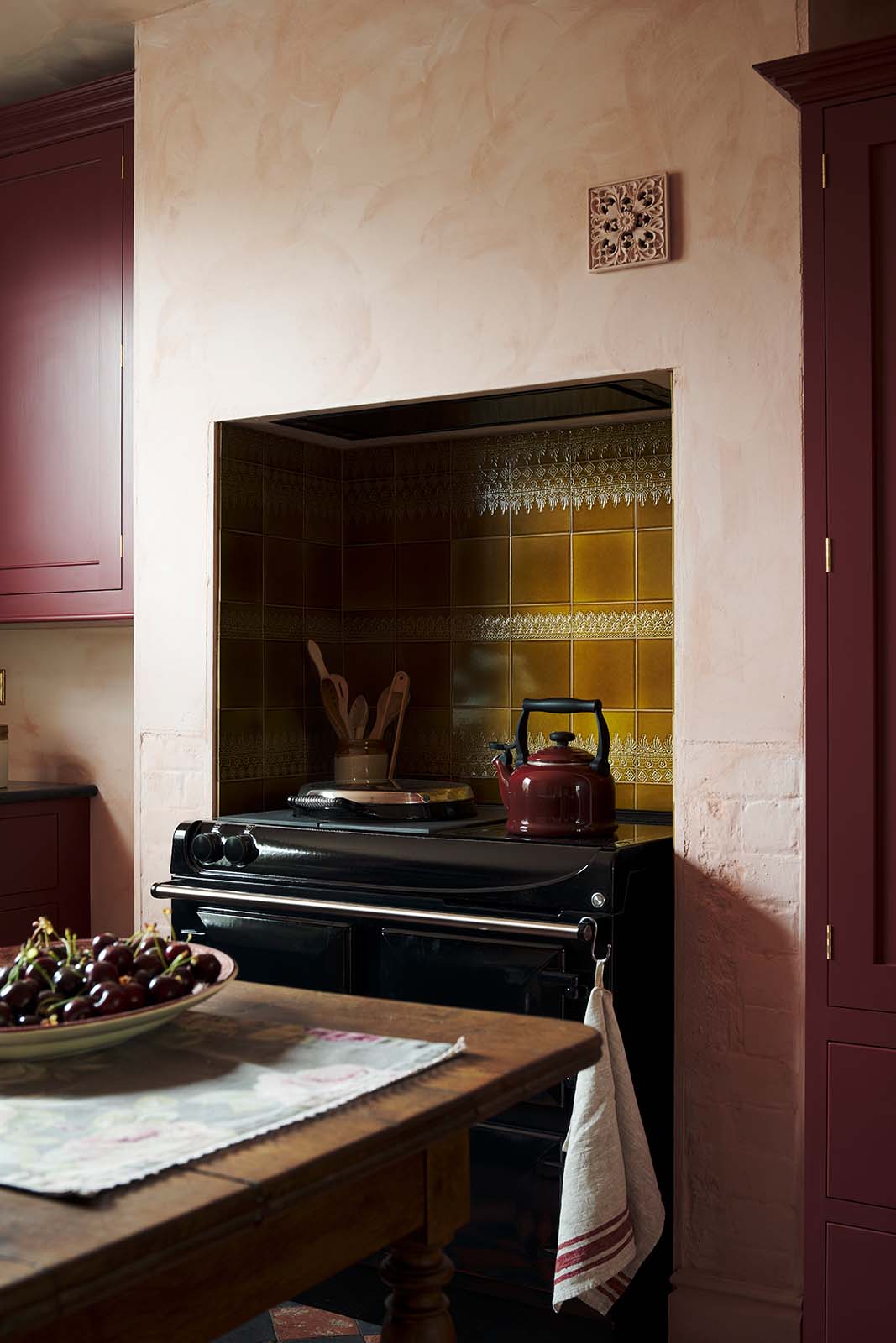 An oblique view of a warm and simply gorgeous refectory-red Shaker kitchen features a sink run with a black granite worktop, a wall cupboard, a pantry cupboard, and a gas stove in a chimney breast with Sepia Lace market splashback tiles. Beautifully decorated with vintage furniture, artisan ceramics, and a matching red teapot, it creates a humble and endearing space.