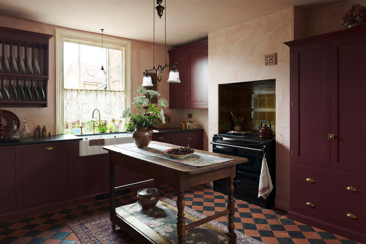 An oblique view of a warm and simply gorgeous refectory-red Shaker kitchen features a sink run with a black granite worktop, a wall cupboard, a small Heirloom plate rack, a pantry cupboard, and a gas stove in a chimney breast with Sepia Lace market splashback tiles. Beautifully decorated with vintage furniture, artisan ceramics, original botanical illustrations, reeded glass pendant lights, and a wild floral arrangement, it creates a humble and endearing space.