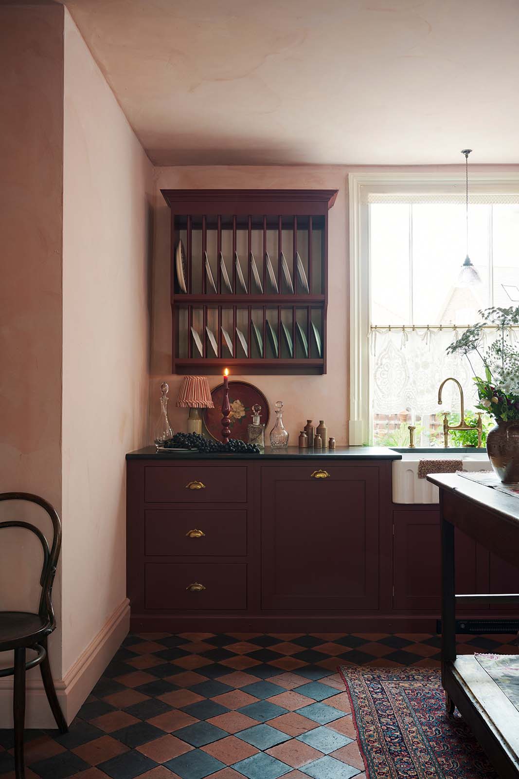 A warm and simply gorgeous refectory-red Shaker kitchen features a sink run with a black granite worktop, an aged brass Ionian tap, a wall cupboard, and a small Heirloom plate rack. Beautifully decorated with vintage furniture, artisan ceramics, original botanical illustrations, and a wild floral arrangement, it creates a humble and endearing space.