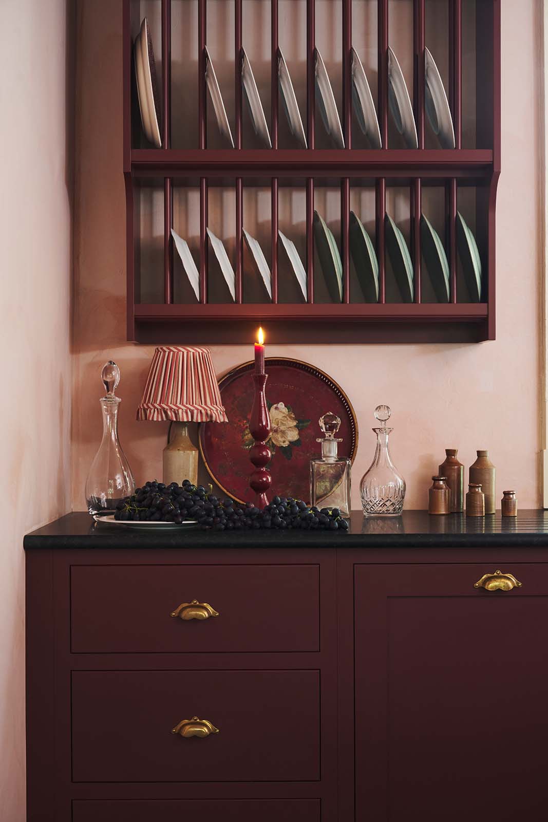 A warm and simply gorgeous refectory-red Shaker kitchen features a sink run with a black granite worktop, an aged brass Ionian tap, a wall cupboard, and a small Heirloom plate rack. Beautifully decorated with vintage furniture, artisan ceramics, original botanical illustrations, and a wild floral arrangement, it creates a humble and endearing space.