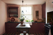 A warm and simply gorgeous refectory-red Shaker kitchen features a sink run with a black granite worktop, a wall cupboard, a small Heirloom plate rack, a pantry cupboard, and a gas stove in a chimney breast with Sepia Lace market splashback tiles. Beautifully decorated with vintage furniture, artisan ceramics, original botanical illustrations, reeded glass pendant lights, and a wild floral arrangement, it creates a humble and endearing space.