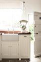 Minimalist white linen Shaker kitchen with a dark sapele wood worktop, Belfast sink, and antique silver taps. Bright light pours in from the garden door, highlighting vintage scales with green pears.