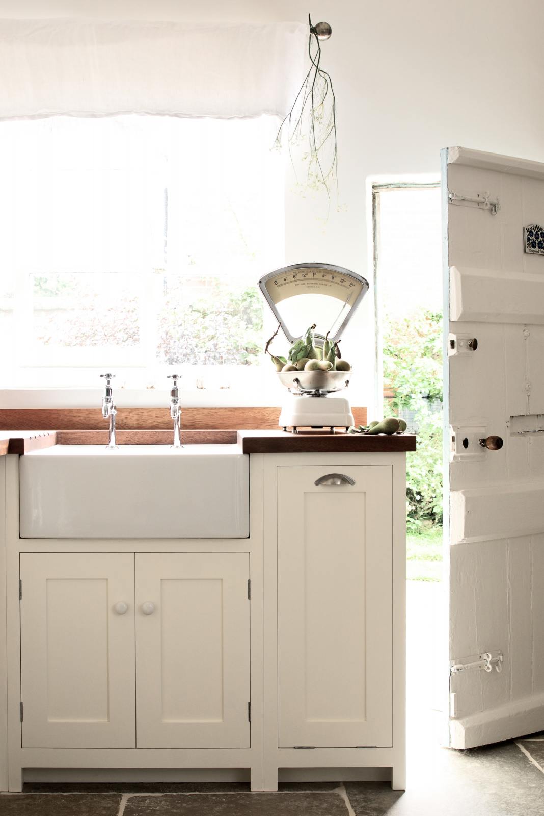 Minimalist white linen Shaker kitchen with a dark sapele wood worktop, Belfast sink, and antique silver taps. Bright light pours in from the garden door, highlighting vintage scales with green pears.