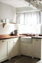 A minimalist white linen Shaker kitchen with a dark polished sapele worktop, countertop cupboard, and L-shaped sink run. Styled with simple touches—wildflowers in glasses, a cylinder bread bin, frames, and mugs on open shelves.