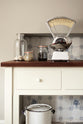 A close-up of a freestanding white linen prep table with a dark polished sapele worktop, styled with glass jars of ingredients, garlic in a glass, and a vintage scale holding chillies. The unused fireplace is finished with Delft-style ceramic splashback tiles.