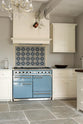 A cooker run in a minimalist white linen Shaker kitchen with a Carrara marble worktop, matching cupboard, and Shaker canopy. A muted blue gas cooker sits beneath a detailed blue floral splashback.