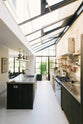 A high view of a fresh and calming Sebastian Cox kitchen features a long cooker run with a cement worktop and splashback, a kitchen island with a sink, a full-height larder cupboard, and a stainless steel canopy. Adorned with a bespoke moss green colour tint, copper pans, artisan ceramics, dried ingredients in glass jars, and books, this kitchen creates a practical earth-toned space that complements the original limestone walls.