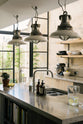 A close-up and oblique view of a fresh and calming Sebastian Cox kitchen features a long cooker run with a cement worktop and splashback, and a kitchen island with a sink. Adorned with a bespoke moss green colour tint, industrial pendant lights, artisan ceramics, pears, bananas, and a jug of fresh lemon water, this kitchen creates a practical earth-toned space that complements the original limestone walls.