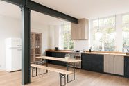 An angled view of a simple and contemporary Sebastian Cox kitchen features a long sink and cooker run with a solid oak worktop, a canopy, and a glazed freestanding cupboard. Adorned with bare pendant lights, an Oktoberfest bench and table, colourful ceramics, and a white refrigerator, this kitchen creates a cosy space with industrial details and a practical finish.
