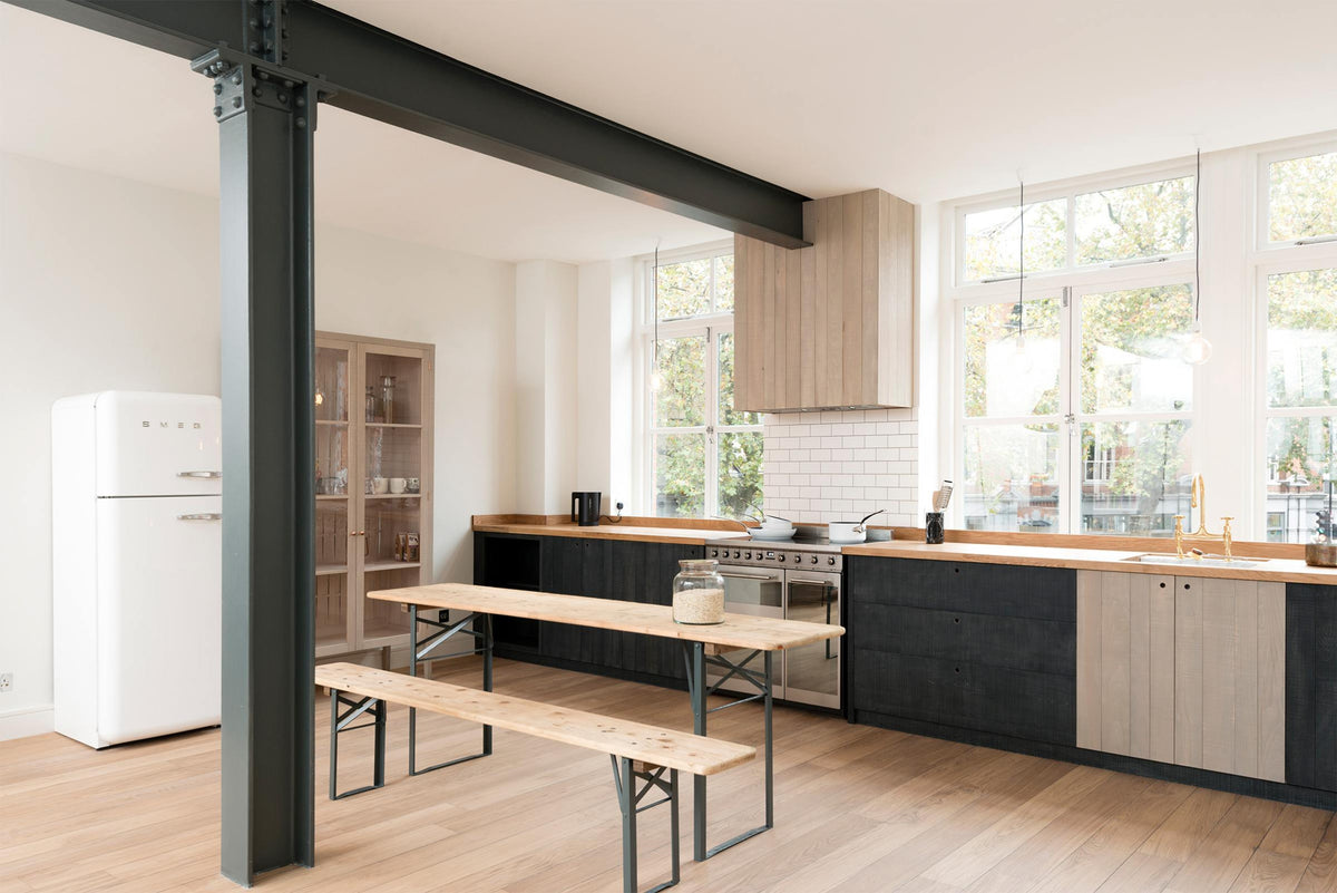 An angled view of a simple and contemporary Sebastian Cox kitchen features a long sink and cooker run with a solid oak worktop, a canopy, and a glazed freestanding cupboard. Adorned with bare pendant lights, an Oktoberfest bench and table, colourful ceramics, and a white refrigerator, this kitchen creates a cosy space with industrial details and a practical finish.