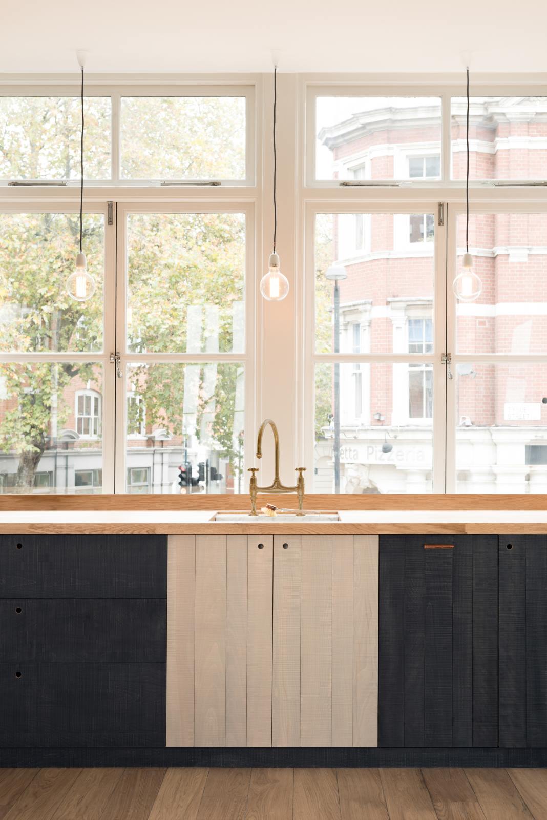 A simple and contemporary Sebastian Cox kitchen features a long sink run with a solid oak worktop, a single Belfast sink, and an aged brass Ionian tap. Adorned with bare pendant lights, a wooden cleaning brush, and a beautiful view of East London, this kitchen creates a cosy space with industrial details and a practical finish.