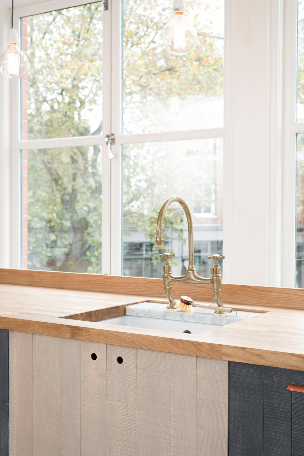 An oblique view of a simple and contemporary Sebastian Cox kitchen features a long sink run with a solid oak worktop, a single Belfast sink, and an aged brass Ionian tap. Adorned with bare pendant lights, a wooden cleaning brush, and a beautiful view of East London, this kitchen creates a cosy space with industrial details and a practical finish.