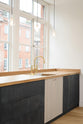 An oblique view of a simple and contemporary Sebastian Cox kitchen features a long sink, cooker run, a canopy, and a glazed freestanding cupboard. Adorned with bare pendant lights, an Oktoberfest bench and table, colourful ceramics, soft pillows, a metal beam structure, and a white refrigerator, this kitchen creates a cosy space with industrial details and a practical finish.