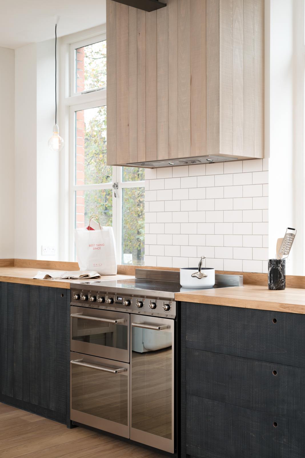 An oblique view of a simple and contemporary Sebastian Cox kitchen’s cooker run with a canopy, and a solid oak worktop. Adorned with bare pendant lights, a silver electrical hob, a black marble utensil holder, and a white refrigerator, this kitchen creates a cosy space with industrial details and a practical finish.
