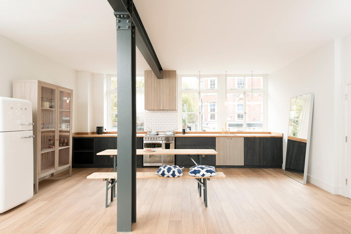 A simple and contemporary Sebastian Cox kitchen features a long sink and cooker run with a solid oak worktop, a single Belfast sink, an aged brass Ionian tap, a canopy, and a glazed freestanding cupboard. Adorned with bare pendant lights, an Oktoberfest bench and table, comfy pillows, a large mirror, and a white refrigerator, this kitchen creates a cosy space with industrial details and a practical finish.
