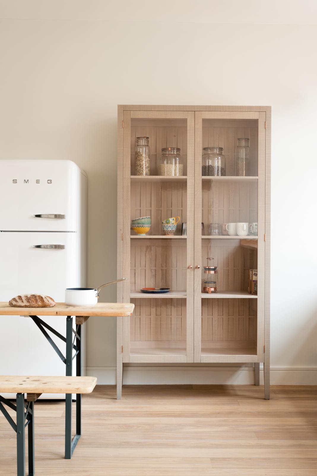 A simple and contemporary Sebastian Cox kitchen features a glazed freestanding cupboard. Adorned with colourful ceramics, a white pan, a fresh loaf of bread, large glass containers of dried ingredients, a collection of tea, a copper coffee press, and a white refrigerator, this kitchen creates a cosy space with industrial details and a practical finish.