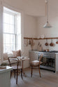 A quiet corner of a warm and natural Sebastian Cox kitchen features a sink and cooker run with a Carrara marble worktop, a single Belfast sink, an aged brass Ionian tap, and a long oak peg rail. Beautifully decorated with vintage furniture, artisan ceramics, a creamware rise-and-fall light, and pale wooden chopping boards, this kitchen creates a calm and delicate atmosphere that celebrates the use of natural materials.
