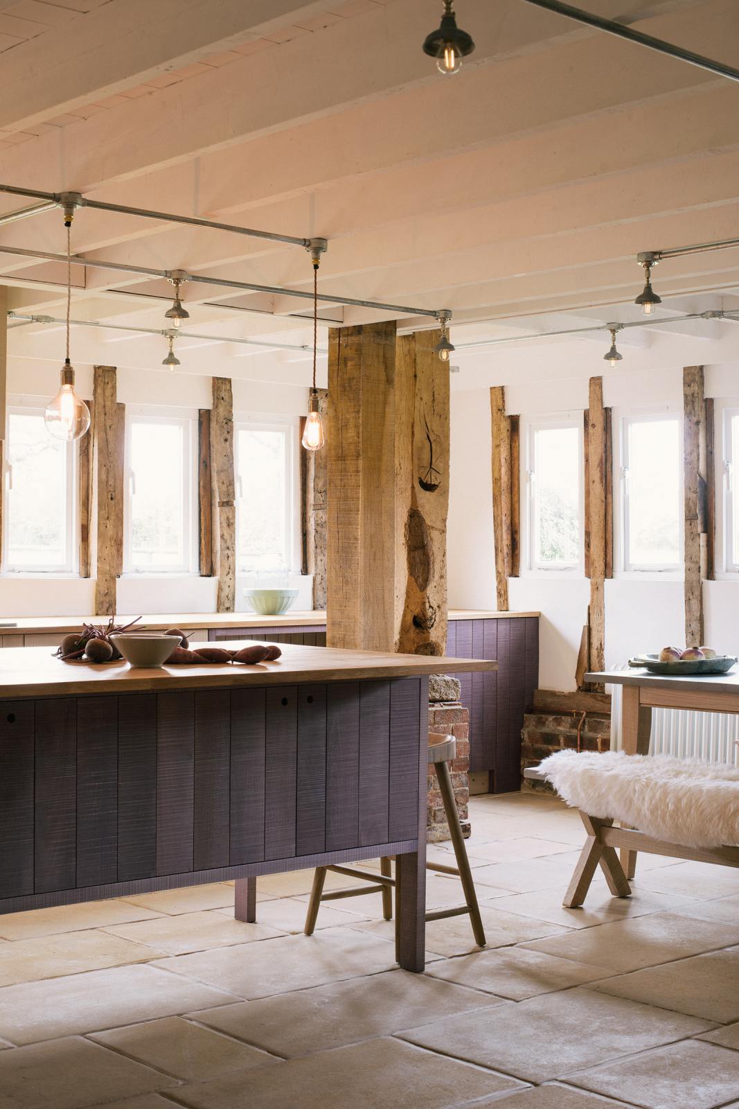 A quiet corner of a soft and spacious Sebastian Cox barn kitchen features a long sink and cooker run with a solid oak worktop, and a kitchen island with a breakfast nook. Beautifully decorated with lavender colour tints, bare pendant lights, vintage furniture, artisan ceramics, sweet potatoes, and sheep rugs, this kitchen creates an elegant and ethereal space.