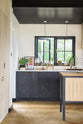 A quiet corner of a large and minimal Sebastian Cox kitchen features a long sink and cooker run with a Carrara marble worktop, a single copper sink, an aged brass Ionian tap, a canopy, a built-in bank of cupboards, and a large kitchen island with a solid oak worktop. Beautifully decorated with artisan ceramics, dried ingredients in glass jars, a water carafe, and drinking glasses, this kitchen creates a simple and practical space.