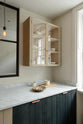 A quiet corner of a sleek and modern Sebastian Cox galley kitchen features a sink run with Carrara marble worktop, a transom partition window, and a glazed wall cupboard. Adorned with simple ceramics, bare pendant lights, a bowl of garlic cloves, and a pale chopping board, this kitchen creates an elegant and practical space.