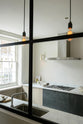 An oblique view past a black-framed transom partition window is a sleek and modern Sebastian Cox galley kitchen featuring a sink and cooker run with a Carrara marble worktop. Adorned with simple ceramics, beaker wall lights, a metal canopy, a chopping board, a rolling pin, and tea towels, this kitchen creates an elegant and practical space.