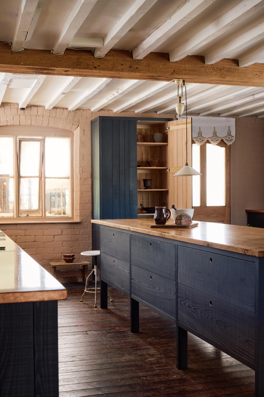 An oblique view of a warm and stylish Sebastian Cox kitchen features a sink run with two single copper sinks, aged brass Mayan taps, glazed partitions, a copper worktop, a prep table with a solid oak worktop, and a freestanding cupboard. Adorned with a creamware rise and fall light, artisan ceramics, and vintage wooden items, this kitchen creates a humble space that celebrates a love of simple and natural materials.