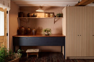 A quiet corner of a warm and stylish Sebastian Cox kitchen features a small prep table with a copper worktop and a bank of free-standing cupboards. Adorned with a milk glass task light, artisan ceramics, and vintage wooden items, this kitchen creates a humble space that celebrates a love of simple and natural materials.