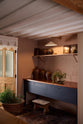 An oblique view of a warm and stylish Sebastian Cox kitchen features a small prep table with a copper worktop and a bank of free-standing cupboards. Adorned with a milk glass task light, artisan ceramics, and vintage wooden items, this kitchen creates a humble space that celebrates a love of simple and natural materials.