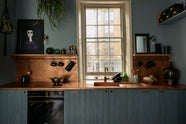 A sweet and simple Clerkenwell blue Sebastian Cox kitchen features a short cooker and sink run with a copper worktop and splashback, a single copper sink, an electric cooker, and shelves with brass hanging rails. Adorned with artisan ceramics, vintage bric-a-brac, aubergines, a sweetheart lettuce, loaves of bread, hanging plants, a framed quirky illustration, and bare wall lights, this kitchen creates a glamorous and practical space.