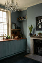 An angled view of a sweet and simple Clerkenwell blue Sebastian Cox kitchen features a short cooker and sink run with a copper worktop and splashback, a single copper sink, an electric stove and shelves with brass hanging rails. Adorned with artisan ceramics, vintage bric-a-brac, aubergines, hanging plants, a framed quirky illustration, a crystal chandelier, and bare wall lights, this kitchen creates a glamorous and practical space.