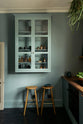 A sweet and simple Clerkenwell blue Sebastian Cox kitchen features a short cooker and sink run with a copper worktop and splashback, and a glazed wall cupboard. Adorned with artisan ceramics, vintage bric-a-brac, hanging plants, a framed quirky illustration, and Helen stools, this kitchen creates a glamorous and practical space.