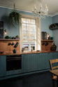 An angled view of a sweet and simple Clerkenwell blue Sebastian Cox kitchen features a short cooker and sink run with a copper worktop and splashback, a single copper sink, an electric stove and shelves with brass hanging rails. Adorned with artisan ceramics, vintage bric-a-brac, a beautiful emerald green teapot, hanging plants, a crystal chandelier, and bare wall lights, this kitchen creates a glamorous and practical space.