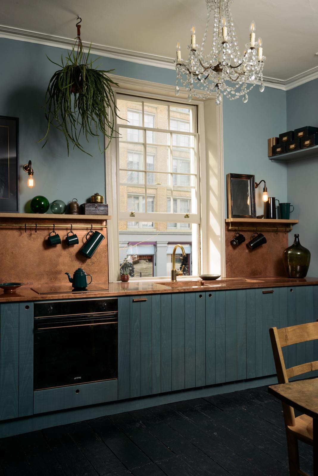 An angled view of a sweet and simple Clerkenwell blue Sebastian Cox kitchen features a short cooker and sink run with a copper worktop and splashback, a single copper sink, an electric stove and shelves with brass hanging rails. Adorned with artisan ceramics, vintage bric-a-brac, a beautiful emerald green teapot, hanging plants, a crystal chandelier, and bare wall lights, this kitchen creates a glamorous and practical space.