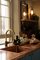 A close-up of a sweet and simple Clerkenwell blue Sebastian Cox kitchen features a short cooker and sink run with a copper worktop and splashback, a single copper sink, an electric stove, and shelves with brass hanging rails. Adorned with artisan ceramics, vintage bric-a-brac, aubergines, and a bare wall light, this kitchen creates a glamorous and practical space.