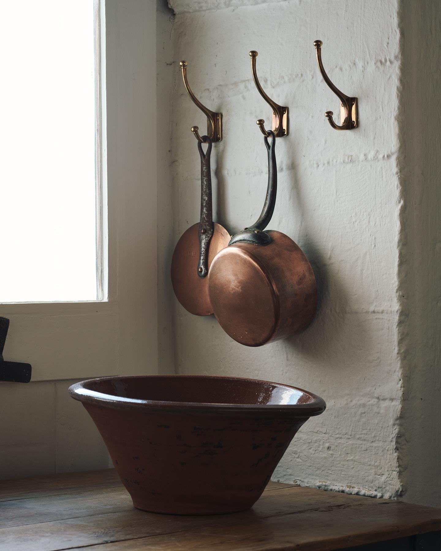 Three Edwardian-style brass hooks, fitted to a white painted wall, keeping up some brass cooking pans.